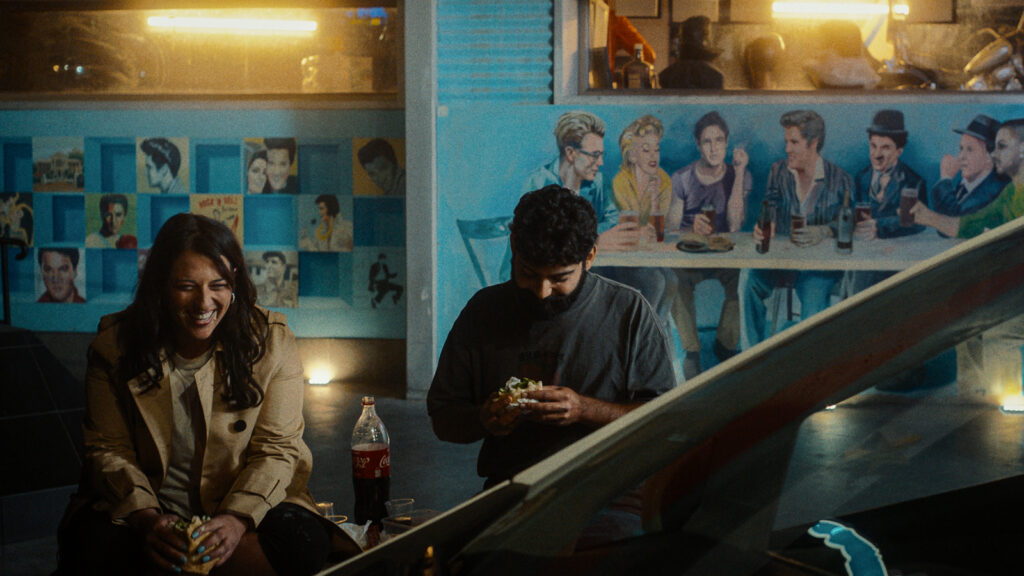Two people sitting and eating food indoors, wall with painted figures behind them