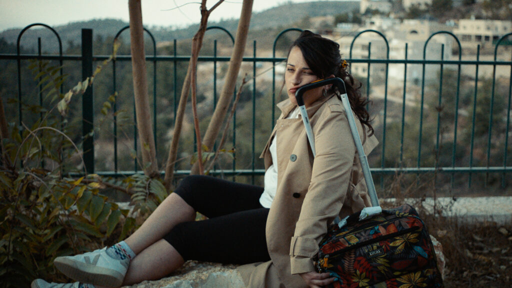 Young woman sitting outdoors beside fence with suitcase, looking to the side