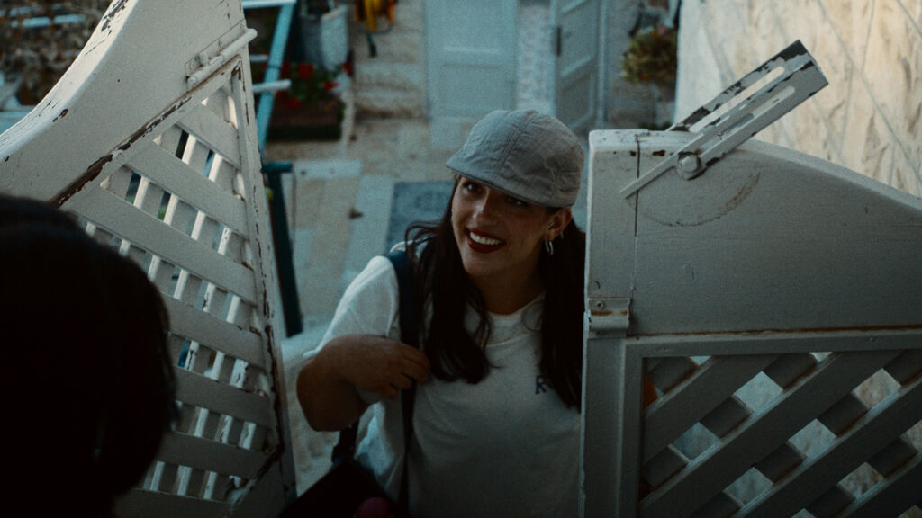 Young woman standing at open gate, leaning forward and smiling toward someone off frame